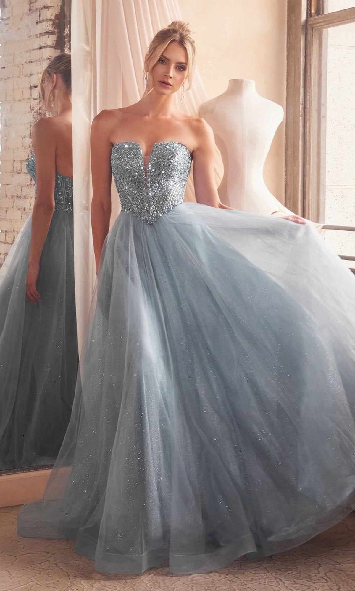 A woman in the Lace-Up Strapless Long Glitter Prom Dress CD0217 stands beside a dress form, holding the skirt of her sparkly blue gown with a large window and mirror in the background.